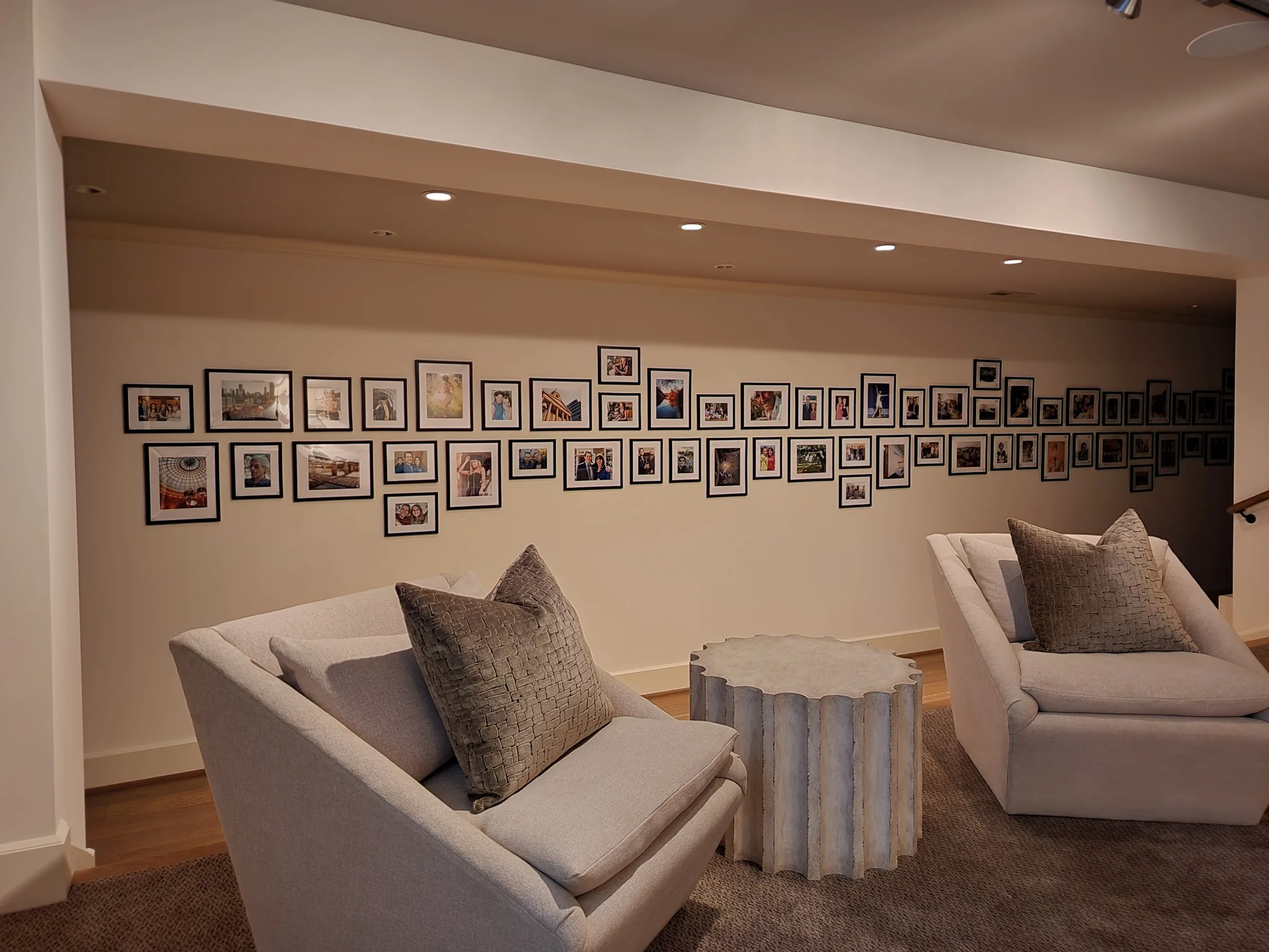 The basement wall: two long courses of black-framed, white-matted photographs running the length of the room above a pair of armchairs.