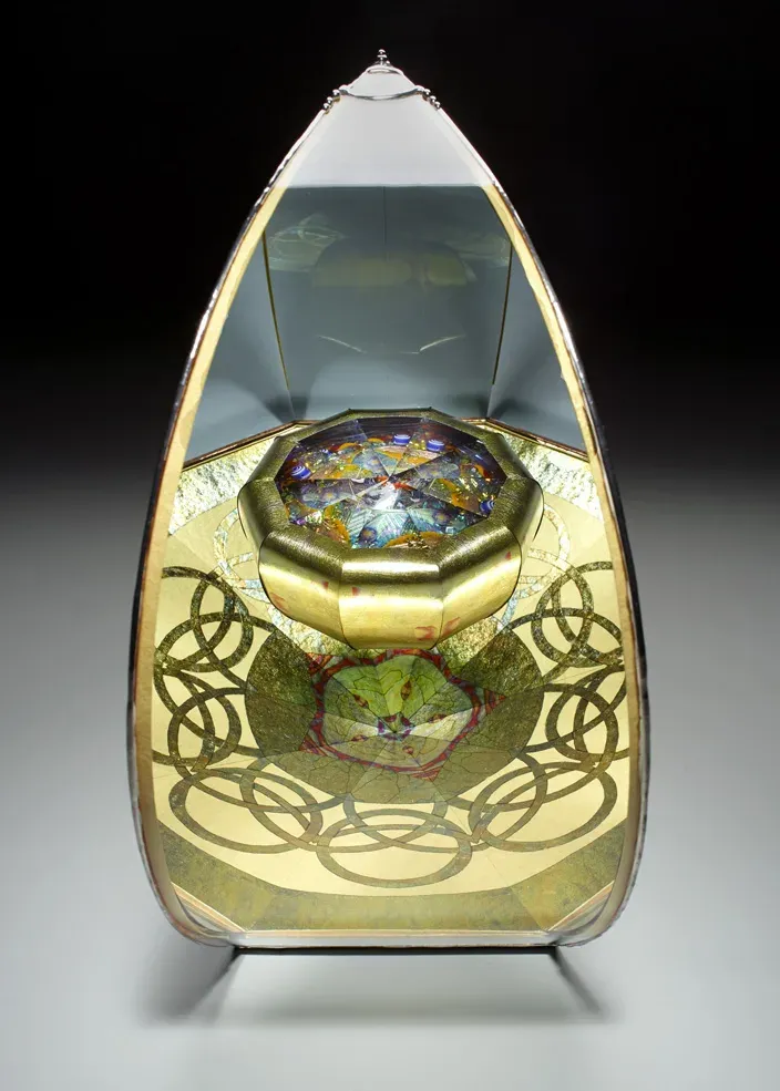 A teardrop-shaped glass kaleidoscope: gold mica reverse-painted panels ringed with interlocking circles, a faceted brass eyepiece chamber above, lit from within.